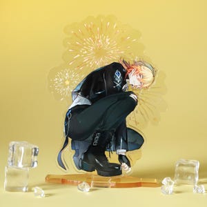 May include: A clear acrylic standee featuring a crouching anime-style character with blonde hair, wearing a black jacket, pants, and boots. The standee has a yellow base and a yellow background with a firework design.