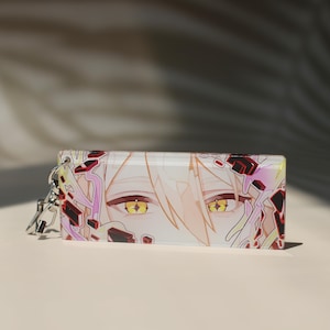 Puede incluir: Un llavero rectangular de acrílico con una ilustración detallada de estilo anime de un personaje con cabello pálido y ojos amarillos brillantes. El diseño incluye formas geométricas abstractas en negro y rosa alrededor de los ojos, unido a un cierre de metal plateado.