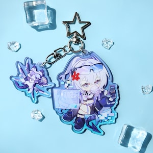Puede incluir: Un llavero acrílico transparente con un personaje de anime de dibujos animados con cabello blanco, vistiendo un atuendo futurista. El llavero tiene un cierre en forma de estrella y un dije más pequeño. El fondo es azul claro con cubitos de hielo.