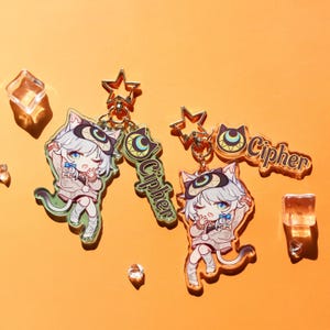 Könnte beinhalten: Zwei Schlüsselanhänger im Anime-Stil mit einem Charakter mit Katzenohren in einem grauen Outfit, mit dem Wort "Cipher" auf einem separaten Anhänger. Die Schlüsselanhänger haben goldfarbene sternförmige Verschlüsse und sind vor einem orangefarbenen Hintergrund mit transparenten, würfelförmigen Akzenten angeordnet.