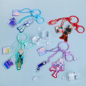 Può includere: Una collezione di portachiavi colorati in stile anime con vari design di personaggi. Ogni portachiavi presenta una figura di personaggio, una piccola immagine quadrata e un ciondolo, tutti collegati da una catena a un portachiavi colorato. I portachiavi sono blu, rossi, verdi e viola.