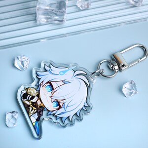 May include: A clear acrylic keychain featuring a cartoon character with white hair and blue eyes. The character is wearing a gold and black outfit. The keychain has a silver clasp and a silver ring. The background is light blue with clear ice-like shapes.