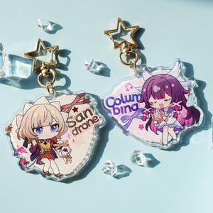 May include: Two anime-style keyrings with star-shaped gold clasps. One keyring features a blonde character with the text "San drone", and the other has a purple-haired character with the text "Columbina". Both keyrings have a clear, decorative border.