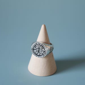 May include: A silver-toned ring with an engraved design, featuring the text "LORD OF THE MYSTERIES". The ring is displayed on a beige, cone-shaped stand against a light blue background. The ring's design includes a central emblem and decorative patterns.
