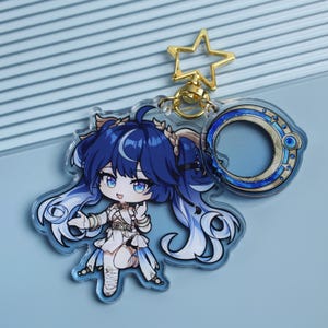 May include: A clear acrylic keyring featuring a cartoon character with blue hair and a white outfit. The keyring has a gold star-shaped clasp and a circular charm with blue and gold accents. The character has large blue eyes and is in a dynamic pose.