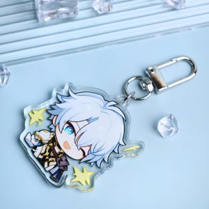 May include: A clear acrylic keychain featuring a cartoon character with white hair and blue eyes. The character is wearing a detailed outfit with gold and navy accents. The keychain has a silver clasp and yellow star accents.