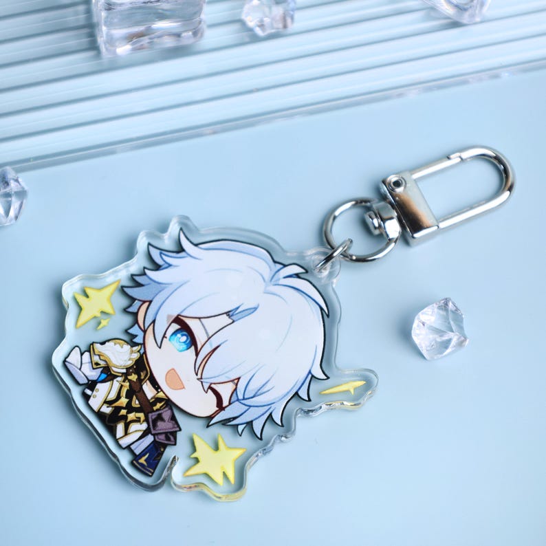 May include: A clear acrylic keychain featuring a cartoon character with white hair and blue eyes. The character is wearing a detailed outfit with gold and navy accents. The keychain has a silver clasp and yellow star accents.