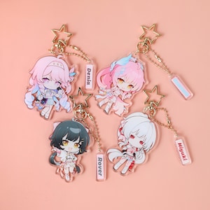 Könnte beinhalten: Vier Acryl-Schlüsselanhänger mit Anime-Figuren, goldenen sternförmigen Verschlüssen und Kugelkettchen. Die Figuren haben rosa, schwarzes und weißes Haar. Kleine rechteckige Anhänger sind mit den Namen Denia, Rover und Hiyuki beschriftet.