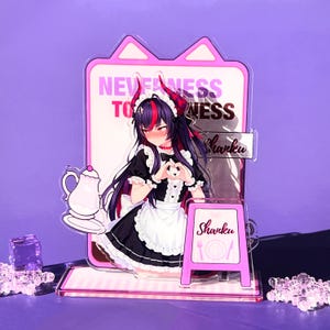 Puede incluir: Un expositor de acrílico con un personaje de estilo anime de pelo oscuro y cuernos, vestido con un traje de sirvienta blanco y negro. El panel muestra el texto NEVERNESS TO NESS y Shanku, junto a un pequeño cartel rosa con el nombre Shanku.