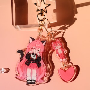 May include: An acrylic keychain featuring a character with long pink hair, cat ears, and glasses, wearing a black jacket and pink skirt. Attached are a smaller pink cat charm and a pink heart charm, all connected by a gold star-shaped clasp.