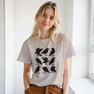 May include: A pale t-shirt displaying a grid of nine black crow illustrations. The crows are shown in different poses, some flying and others standing. The shirt has short sleeves and a round neckline.