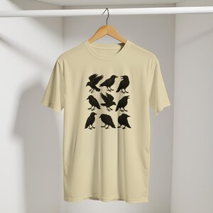 May include: A cream-coloured t-shirt displaying a grid of nine black raven illustrations in different poses. The ravens are shown with detailed wings and beaks. The shirt is hanging on a wooden hanger against a plain background.