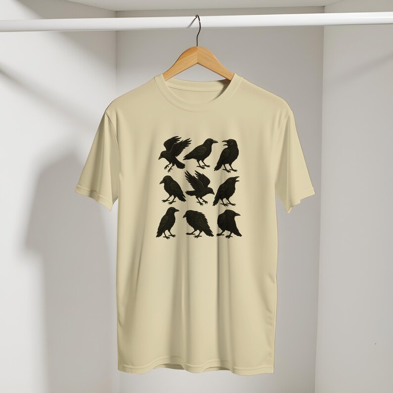 May include: A cream-coloured t-shirt displaying a grid of nine black raven illustrations in different poses. The ravens are shown with detailed wings and beaks. The shirt is hanging on a wooden hanger against a plain background.