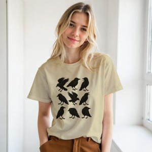 May include: A beige t-shirt with a grid of nine black crow illustrations. The crows are in different poses, some with wings open. The shirt is made of a soft material.