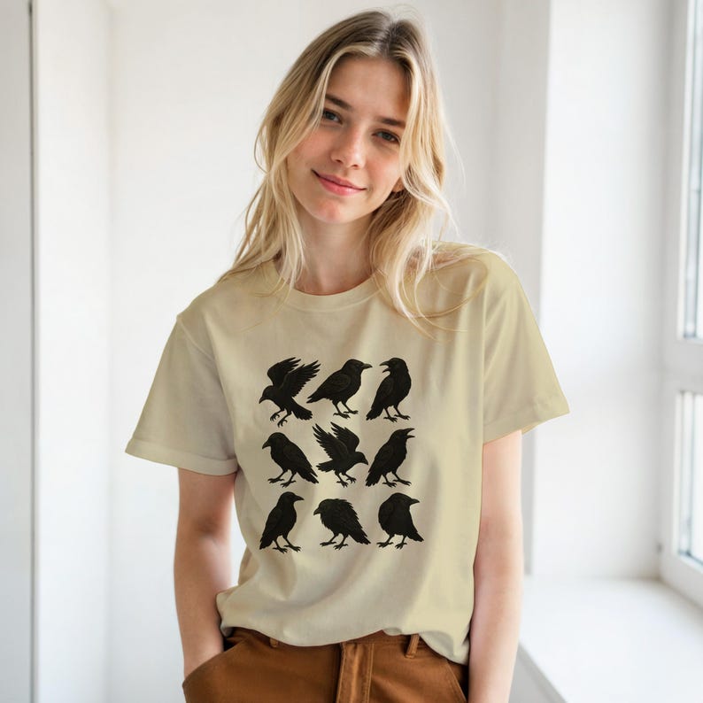 May include: A beige t-shirt with a grid of nine black crow illustrations. The crows are in different poses, some with wings open. The shirt is made of a soft material.