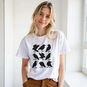 May include: White t-shirt with a grid of nine black raven illustrations. The ravens are shown in different poses, some with wings open, others perched. The shirt is made of a soft material, suitable for casual wear.