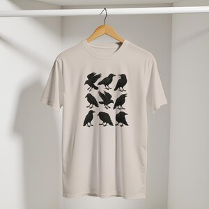 May include: A cream-coloured t-shirt displaying a grid of nine black crow illustrations in different poses. The crows are detailed and set against a neutral background. The shirt is hanging on a wooden hanger.