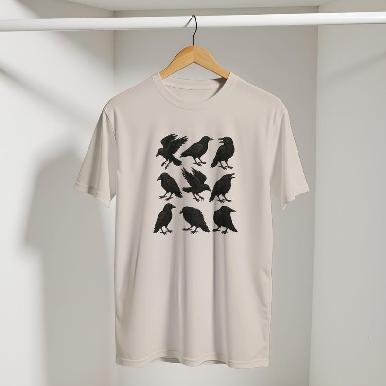 May include: A cream-coloured t-shirt displaying a grid of nine black crow illustrations in different poses. The crows are detailed and set against a neutral background. The shirt is hanging on a wooden hanger.