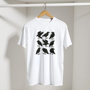 May include: White t-shirt with a design of nine black crows in different positions. The crows are rendered as detailed silhouettes, contrasting sharply against the white material. The shirt is displayed on a wooden hanger.