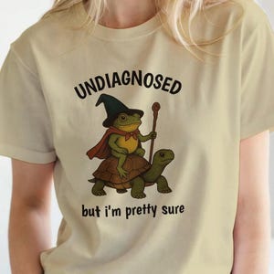 Op de afbeelding: Beige t-shirt met de tekst "UNDIAGNOSED but I'm pretty sure". De afbeelding toont een kikker met een tovenaars hoed en cape die op een schildpad rijdt en een staf vasthoudt.