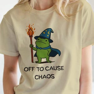 May include: Beige t-shirt with a cartoon frog wizard holding a flaming staff. The frog wears a blue cape and a hat with stars and a crescent moon. The text on the shirt reads "OFF TO CAUSE CHAOS".