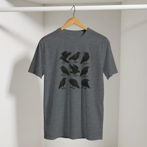 May include: A grey t-shirt displaying a grid of nine black raven illustrations. The ravens are shown in different poses, some with wings extended, others perched. The design is centrally placed on the shirt.