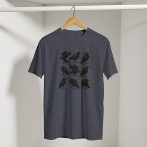 May include: Dark grey t-shirt with a grid of nine black raven illustrations. The ravens are in different poses, some with wings open, others perched. The shirt hangs on a wooden hanger against a white backdrop.