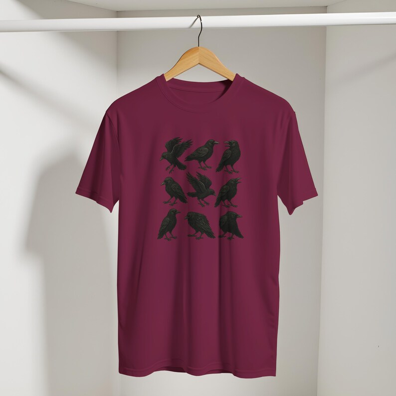 May include: Burgundy t-shirt with a black illustration of nine crows in different poses. The crows are arranged in a three-by-three grid. The shirt is hanging on a wooden hanger.