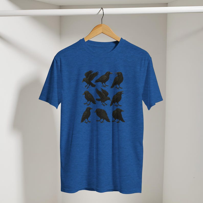 May include: A blue t-shirt with a design of nine black crows. The crows are shown in different positions, with detailed wings and beaks. The shirt is displayed on a wooden hanger. The background is a solid blue colour.