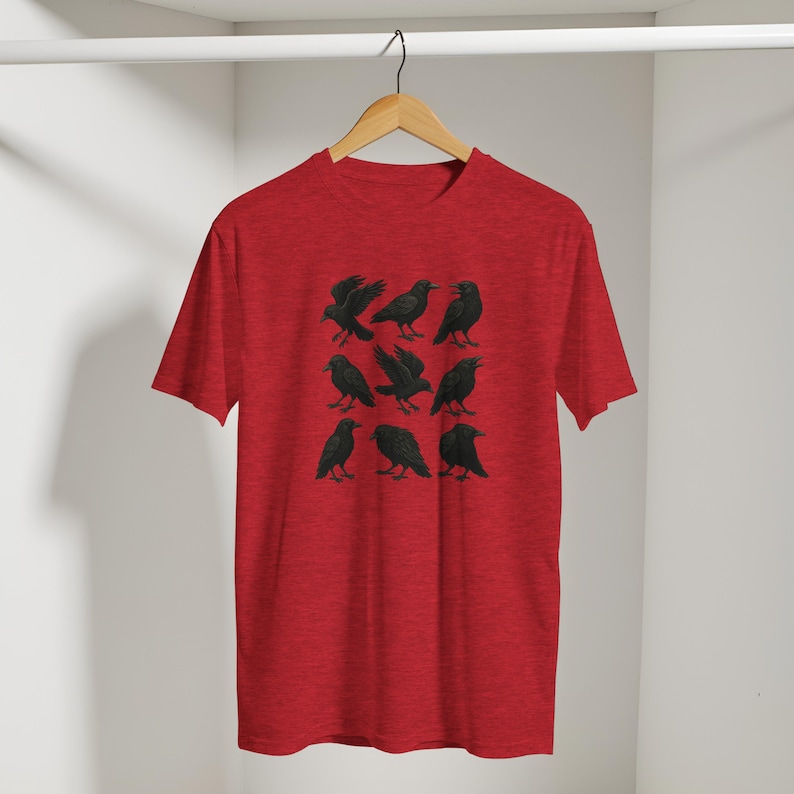 May include: A red t-shirt with a design of nine black raven illustrations arranged in a grid. The ravens are shown in different poses. The t-shirt is hanging on a wooden hanger.