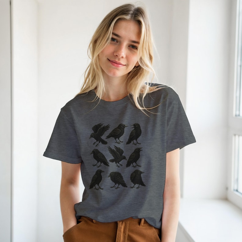 May include: A charcoal grey t-shirt displaying a pattern of nine black crow illustrations. The crows are shown in different positions, some with wings extended. The shirt is made of a soft fabric and has a standard crew neck.