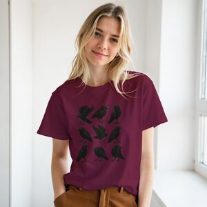May include: A maroon t-shirt with a design of nine black crows. The crows are depicted in different positions, some with outstretched wings. The shirt is made of a soft fabric and has a crew neck.