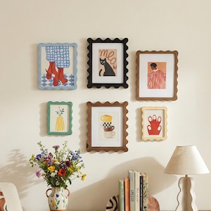 May include: A gallery wall featuring seven framed art prints with scalloped edges. The prints showcase various illustrations, including a cat, a person, and a vase of flowers. The frames are in various colors, including black, brown, green, and blue.