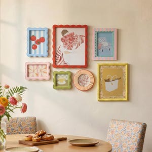 May include: A collection of colorful picture frames with scalloped edges, each containing unique artwork. The frames are in various shapes and sizes, including rectangular and circular, and are arranged on a neutral wall. The artwork includes illustrations of tomatoes, flowers, and a cat in a bag.