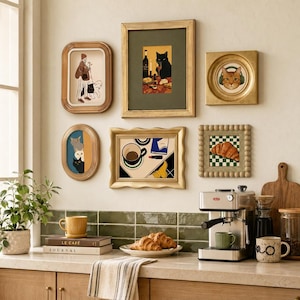 Puede incluir: Una pared de galería con seis láminas enmarcadas que muestran diversas ilustraciones, incluyendo gatos, un cruasán y una taza de café. Debajo, una encimera de cocina contiene una cafetera, libros, un plato con cruasanes y varias tazas.