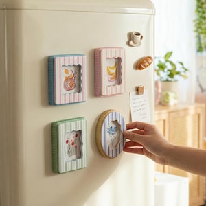 May include: A collection of decorative refrigerator magnets in various shapes and colors. The magnets feature striped frames in blue, pink, and green, with illustrations of fruit, flowers, and a shopping bag. A hand is placing an oval magnet with a floral design.