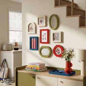 May include: A collection of colorful, decorative picture frames in various shapes and sizes, mounted on a cream-colored wall. The frames are in shades of red, green, blue, and cream, with abstract designs and patterns. A red vase with flowers sits on a wooden cabinet.