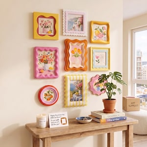 Könnte beinhalten: Eine Galerie-Wand mit neun bunten, wellenförmigen Bilderrahmen, die verschiedene Blumen- und Küstenkunstdrucke enthalten. Darunter steht ein Holzkonsolentisch mit einer Kerze, einer Karte mit Text, einer kleinen Schale und zwei Büchern.