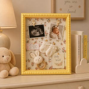 Puede incluir: Un marco de caja amarillo con cuentas y fondo floral. En su interior hay una ecografía, una foto de un bebé, un par de patucos blancos con encaje, un sonajero de conejo de madera y una tarjeta con el texto Hello World y datos del bebé.