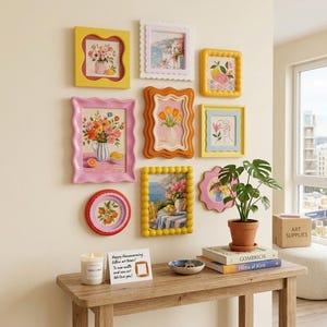 May include: A gallery wall arrangement featuring nine colorful, wavy-edged frames containing various floral and coastal art prints. Below, a wooden console table holds a candle, a greeting card, a small bowl, and a stack of two books.