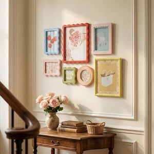 May include: A collection of colorful picture frames with scalloped edges adorns a wall. The frames are in various shapes and sizes, with images of fruit, flowers, and other decorative elements. A wooden table with a vase of roses and books sits below.