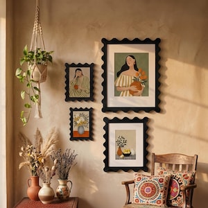 May include: A collection of framed art prints with black scalloped edges, featuring illustrations of women, flowers, and still life arrangements. The artwork is displayed on a beige wall, complemented by a hanging plant and decorative vases.
