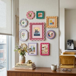 May include: A display of decorative picture frames in assorted colours and designs, hung on a wall. The frames contain images such as a dog, a croissant, and a couple cycling. A vase of flowers is placed on a wooden table beneath the frames.