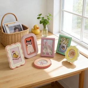 May include: A selection of decorative picture frames in different shapes and sizes, with colourful designs and illustrations. The frames include a cat wearing sunglasses, a bee with the text "Bee Aggressive", and a cat on toast. The frames are displayed on a wooden table.