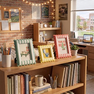 May include: Three scalloped picture frames in light green, yellow, and red, each with white stripes. The frames display illustrations of a ping pong table, a toilet, and a cactus with the text "YOUR ASS KICKIN'" on a wooden shelf with books.