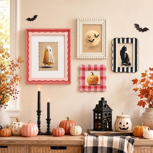 Op de afbeelding: Een Halloween-decoratiecollectie met vier ingelijste kunstwerken van een geest, een maan met vleermuizen, een zwarte kat en een pompoen. De opstelling bevat diverse keramische pompoenen, twee zwarte kandelaars en een zwarte lantaarn.