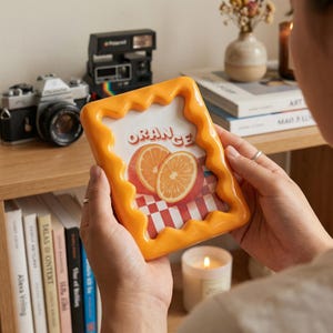 Op de afbeelding: Een rechthoekige fotolijst met een glanzende oranje golvende rand. De lijst bevat een print met twee sinaasappelschijfjes en een rood-wit geblokt patroon met de tekst ORANGE in een vetgedrukt lettertype in het midden bovenaan.