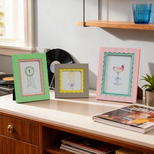 May include: Three colorful picture frames with drawings of a ribbon, a balloon, and a cocktail. The frames are green, gray, and pink, with scalloped edges and pastel accents. The frames are displayed on a white surface.