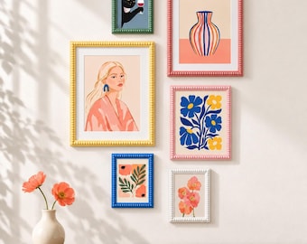 May include: A gallery wall arrangement featuring six framed art prints with beaded frames in green, pink, yellow, and blue. The prints include a black cat with a wine glass, a striped vase, a portrait of a person, and various floral illustrations.