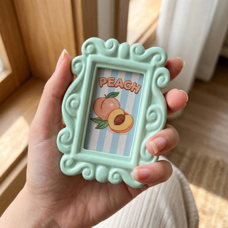 Op de afbeelding: Een klein, licht mintgroen rechthoekig frame met sierlijke, krullende randen. Het midden heeft een verticale lichtblauwe en witte gestreepte achtergrond met een illustratie van een perzik en de tekst PEACH in oranje letters.